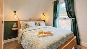 Ein ordentlich bezogenes Bett mit floralem Überwurf, ein Nachttisch mit Lampe und eine Schale mit Croissants und Saft.