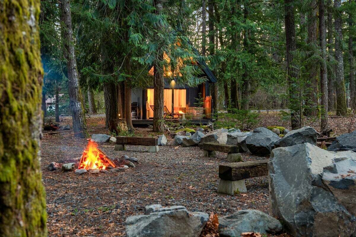 A cabin in a forest with a fire, rocks, and benches.