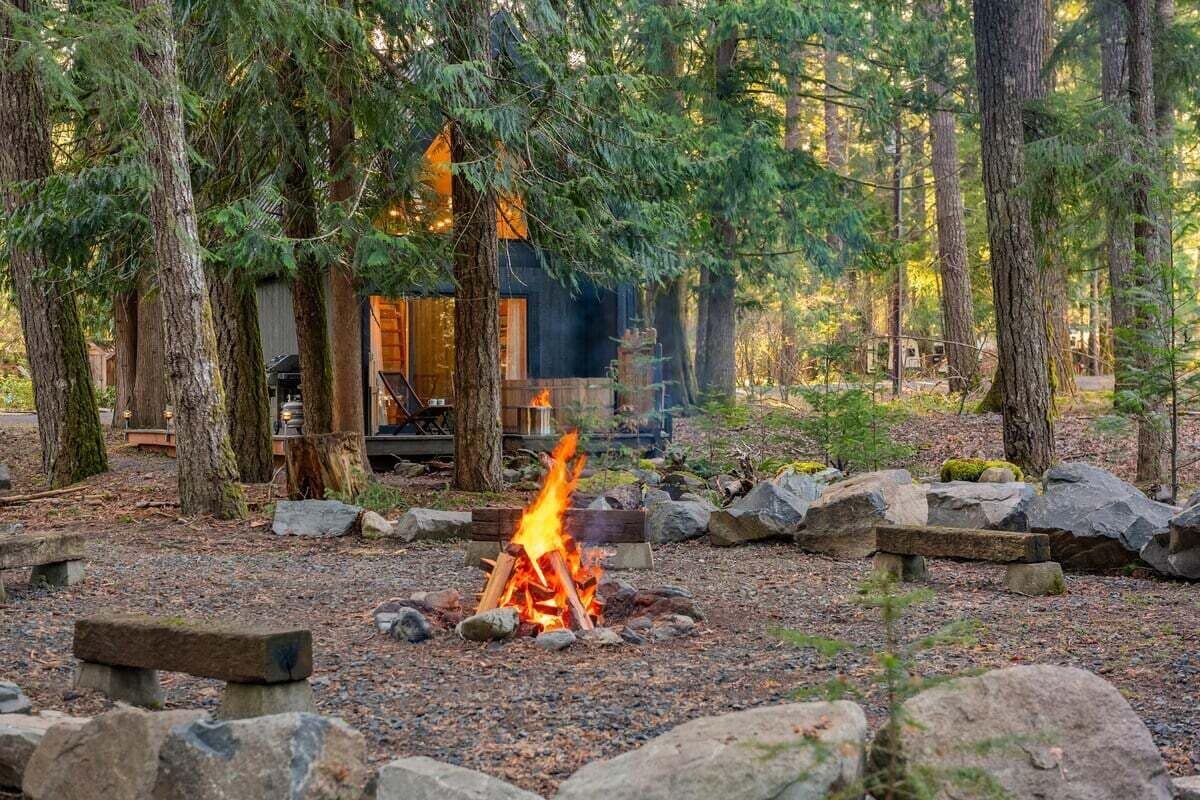 A cabin surrounded by trees with a fire in the foreground.