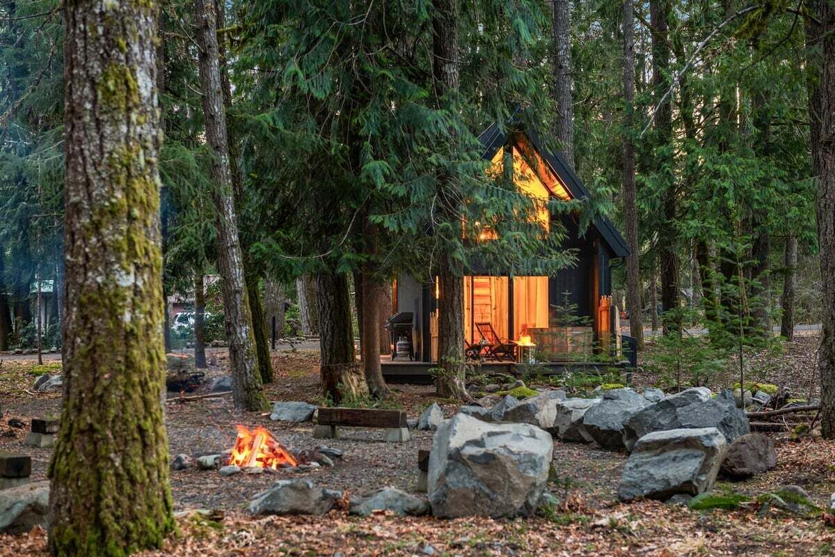 A cabin in a forest with a fire in front, surrounded by rocks and trees.