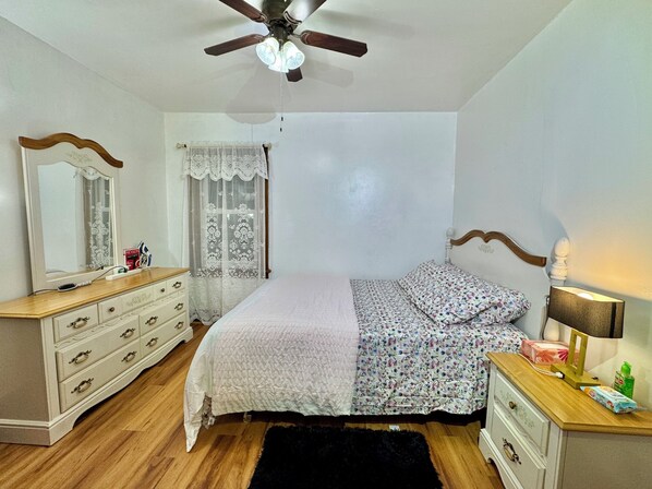 Un dormitorio con cama, cómoda, mesita de noche, ventilador de techo y ventana con cortinas de encaje.