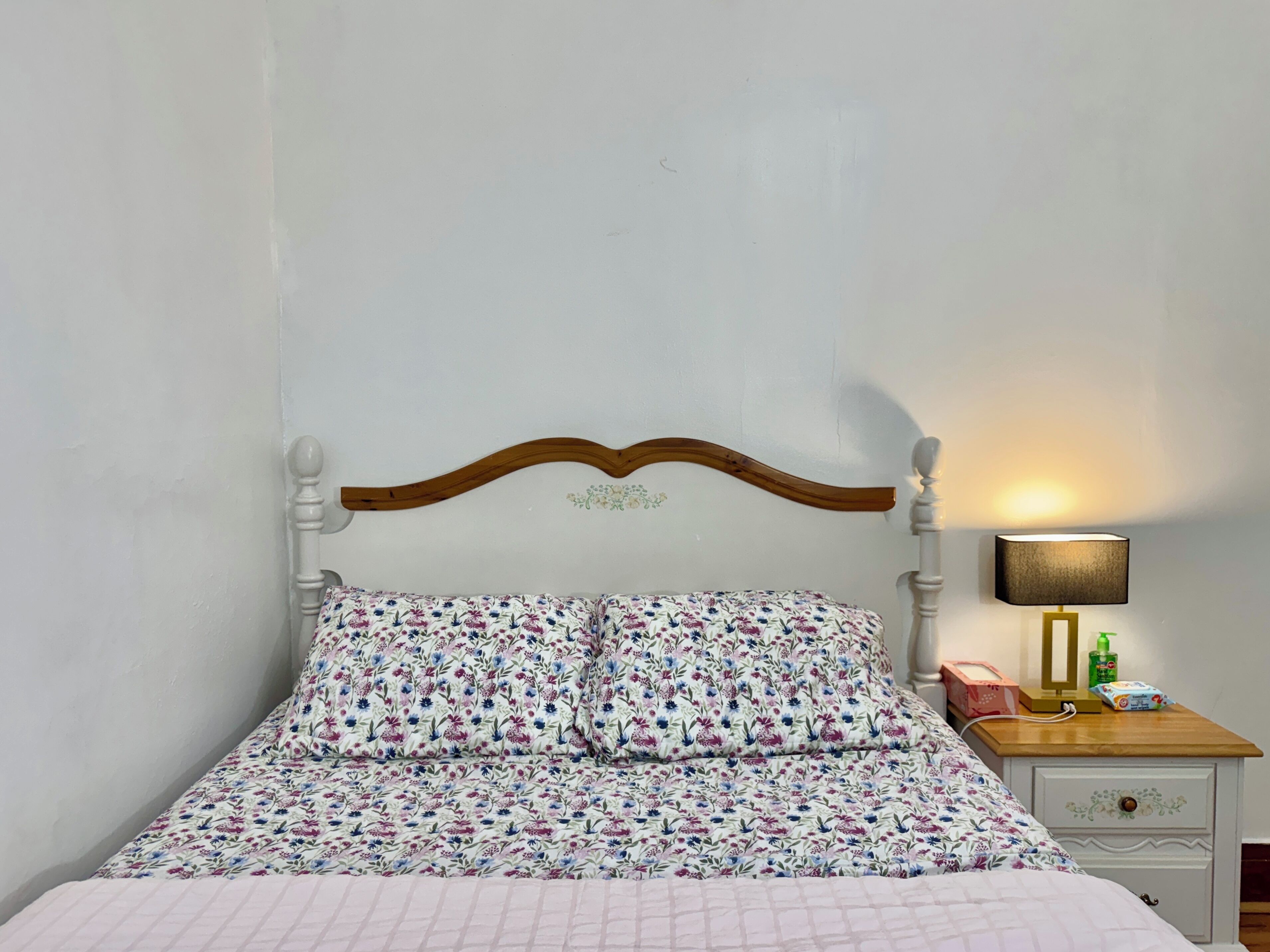 Ein Bett mit floralem Überwurf, ein hölzernes Kopfteil, ein Nachttisch mit Lampe sowie ein Beistelltisch mit Taschentuchbox und Flasche.