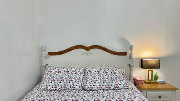 Ein Bett mit floralem Überwurf, ein hölzernes Kopfteil, ein Nachttisch mit Lampe sowie ein Beistelltisch mit Taschentuchbox und Flasche.