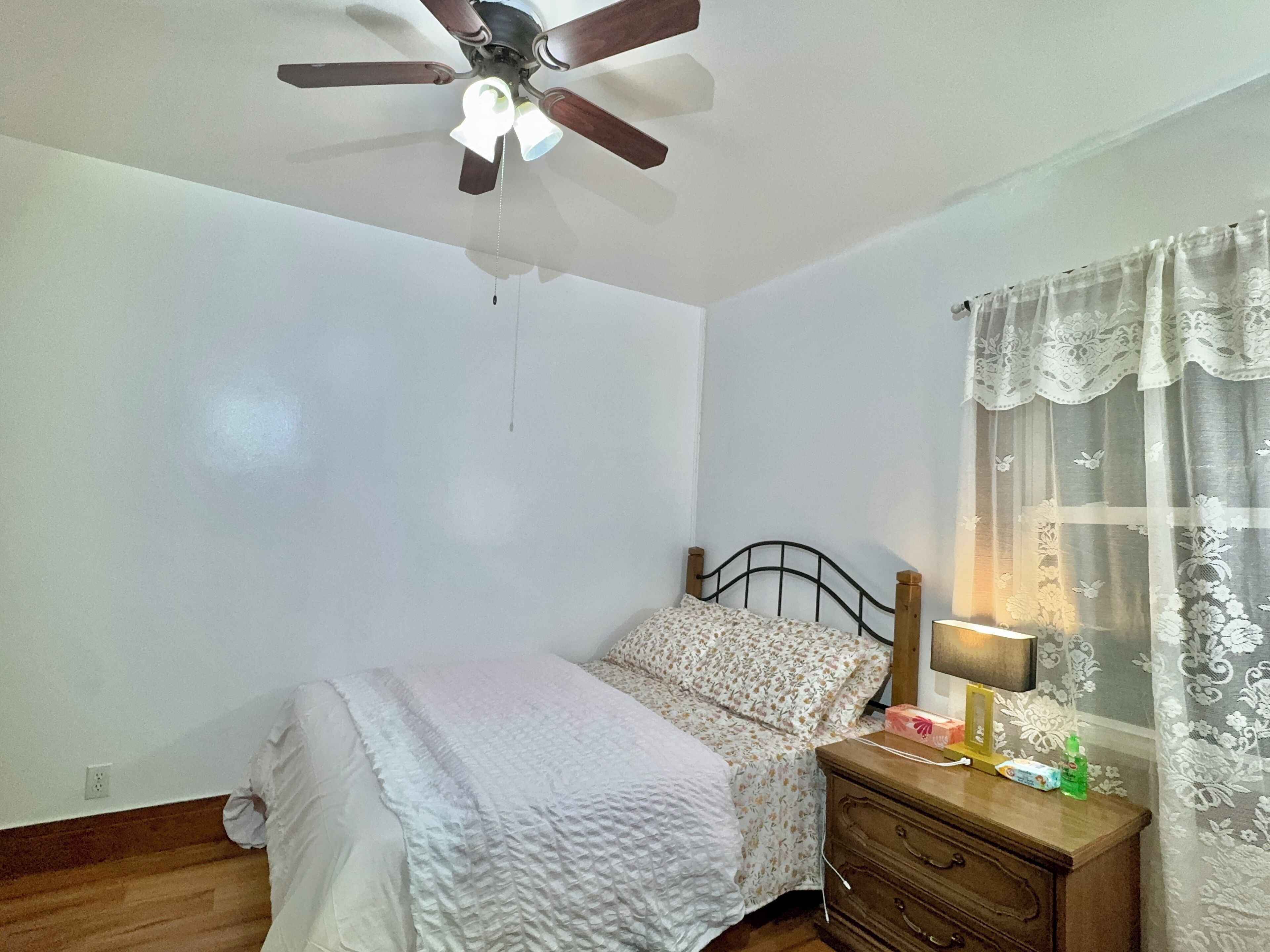 A bedroom with a bed, a wooden dresser, a ceiling fan, and a window with a curtain.
