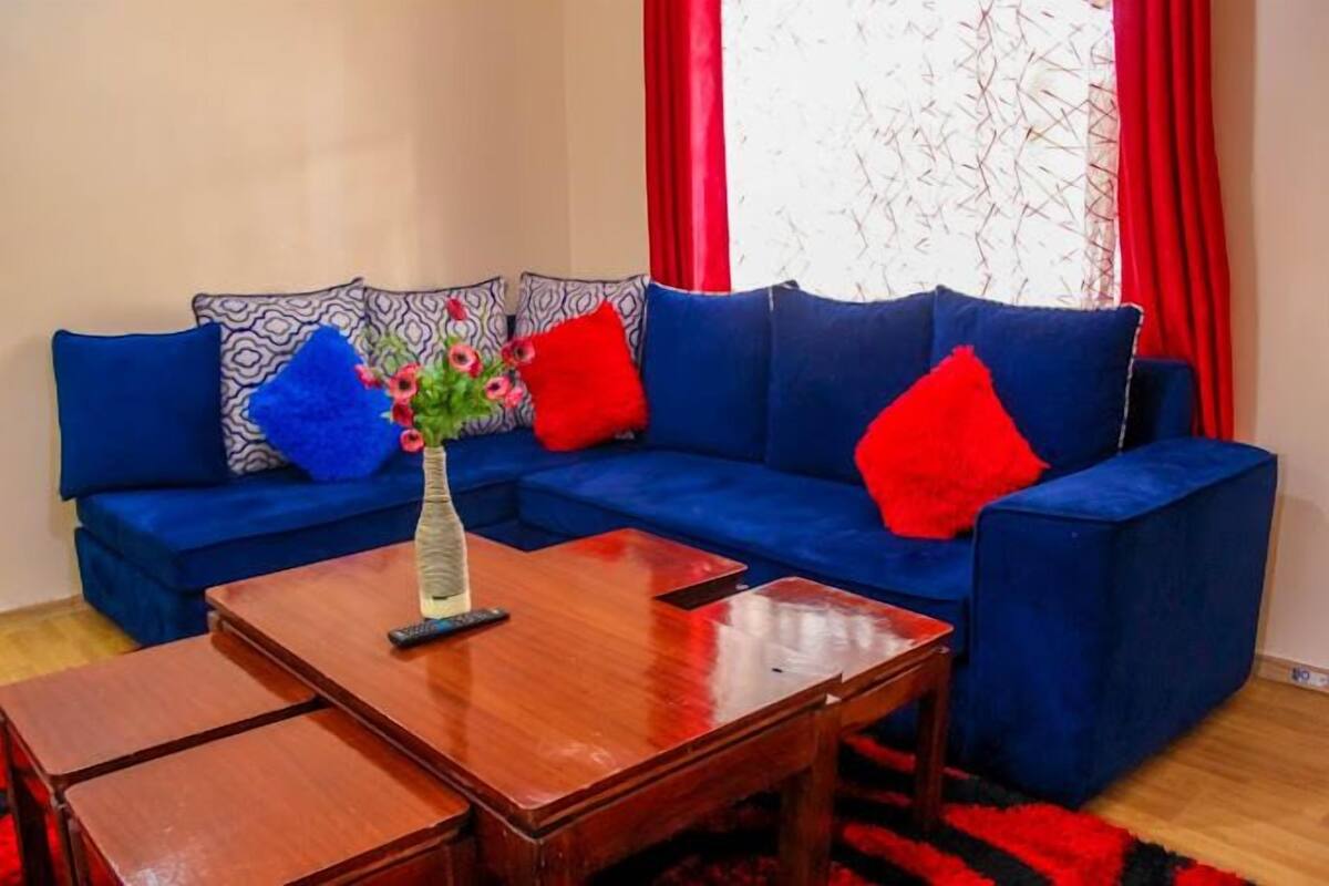 Un sofá azul con cojines rojos y estampados, una mesa de centro de madera, y un jarrón con flores.