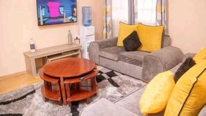 A living room with a grey sofa, yellow cushions, a wooden coffee table, a TV on the wall, and a water dispenser. - Lux Suites Elmoc Apartments Nakuru (Nakuru)