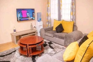 Una sala de estar con sofá gris, cojines amarillos, una mesa de centro de madera, televisor en la pared y dispensador de agua.