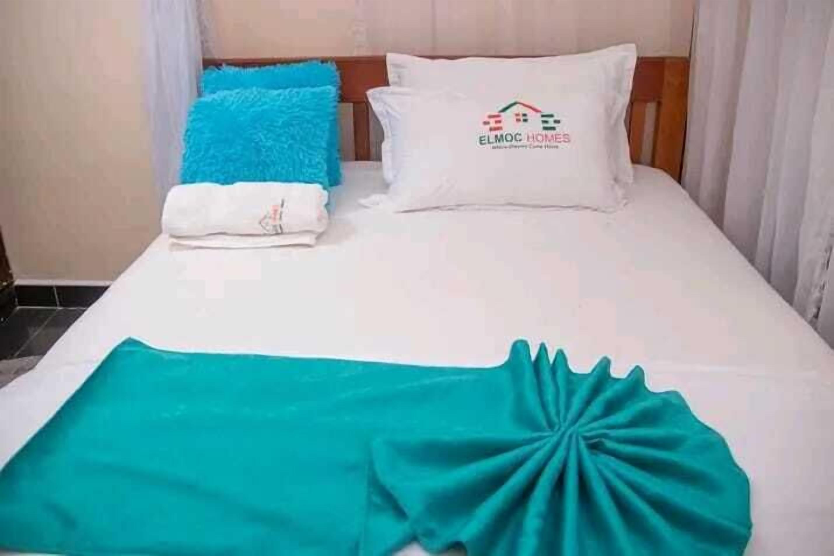 A neatly made bed with a white pillow featuring a logo, a blue pillow, and a teal blanket.