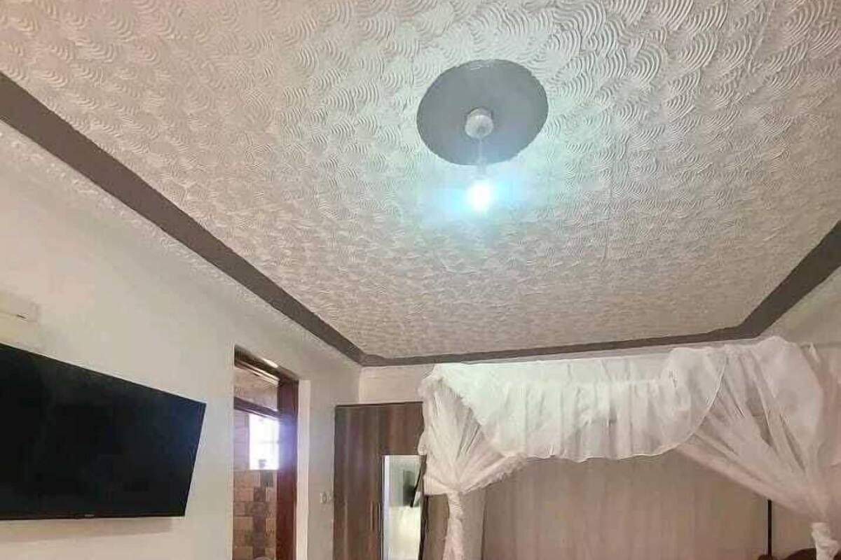 Habitación de hotel con cama bajo dosel, televisor montado en la pared, una pequeña mesa redonda con un libro y un control remoto, y un baño visible a través de una puerta abierta.