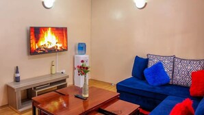 A living room with a flat-screen TV displaying a fireplace, a blue sofa with patterned cushions, a wooden coffee table, and a water dispenser. - Lux Suites Elmoc Apartments Nakuru (Nakuru)