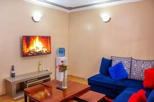 Una sala de estar con una televisión de pantalla plana mostrando una chimenea, un sofá azul con cojines estampados, una mesa de centro de madera y un dispensador de agua.