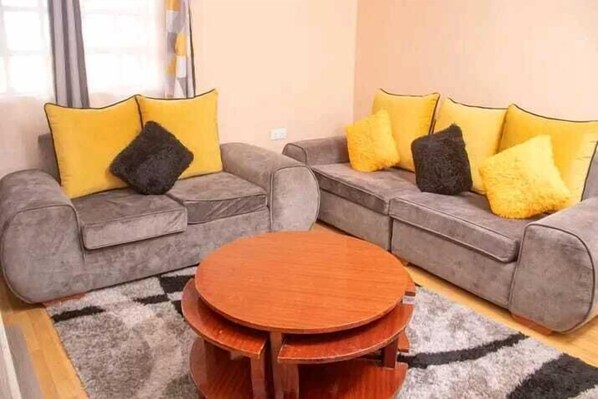 A living room with two grey sofas, each with yellow and black cushions, and a round wooden coffee table. - Lux Suites Elmoc Apartments Nakuru (Nakuru)