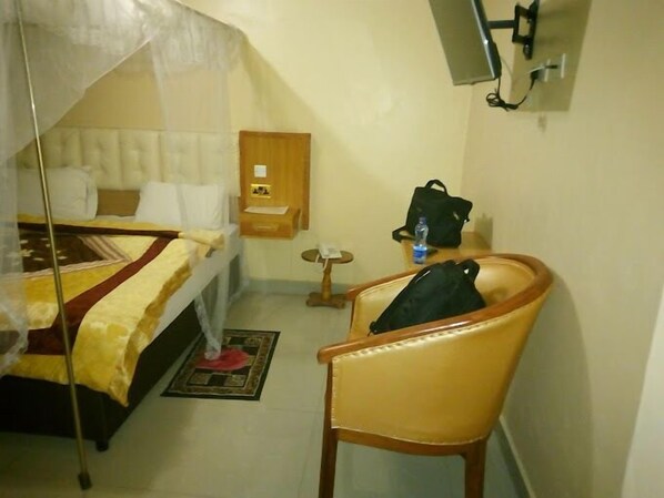 A hotel room with a bed, a chair, a small table, a water bottle, and a wall-mounted TV.