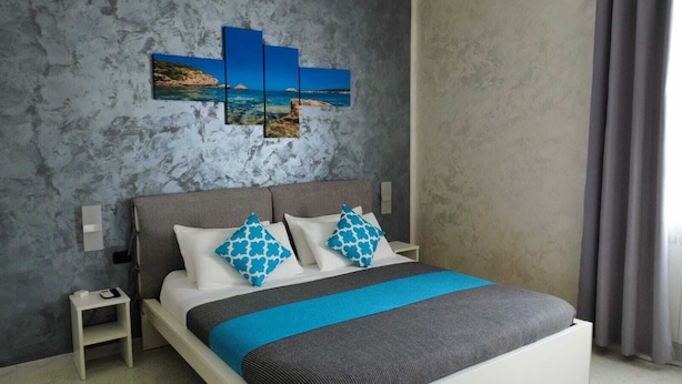 A modern bedroom with a grey and blue color scheme, featuring a bed with a grey and blue bedspread, two matching pillows, a nightstand with a phone and charger, and a wall-mounted artwork depicting a coastal scene. - B&B La Munachell  (Vieste)