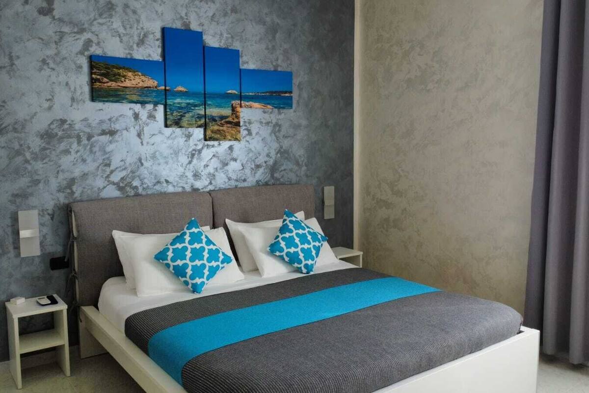 A modern bedroom with a grey and blue color scheme, featuring a bed with a grey and blue bedspread, two matching pillows, a nightstand with a phone and charger, and a wall-mounted artwork depicting a coastal scene.