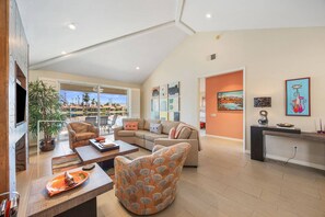 A spacious living area with a large window, a sofa, a coffee table, and a dining table with a plate of food. - Updated Condo on Lake Mirage, Tennis, Pickleball, Pool, Spa, Gorgeous Views (Rancho Mirage)