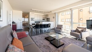 A modern living room with a sofa, coffee table, and chairs, a kitchen with a dining area, and large windows offering a view of the city.