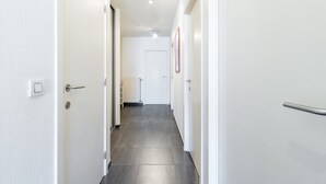 A narrow, well-lit hallway with white walls and a tiled floor, leading to a door at the end.