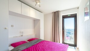 A bedroom with a bed, a nightstand, a window with curtains, and a view of the city. - Apartment in Blankenberge near the Pier (Blankenberge)