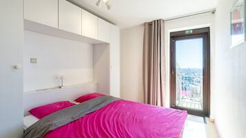 Een slaapkamer met een bed, een nachtkastje, een raam met gordijnen en uitzicht op de stad.