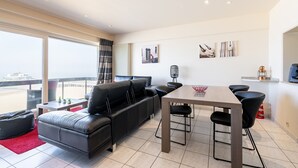 A modern living room with a black leather sofa, a dining table with four chairs, and a large window offering a view of the beach. - Apartment in Blankenberge with Sea View (Blankenberge)