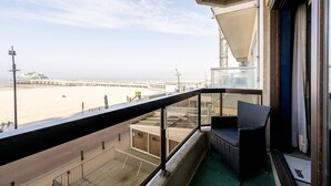 Un balcon avec vue sur la plage et la jetée, une chaise en osier et une rambarde en verre.