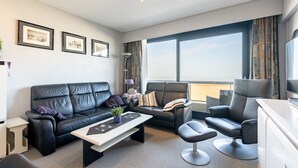 Un salon moderne avec un canapé en cuir noir, un fauteuil assorti et une petite table. Les murs sont ornés de tableaux encadrés, il y a une lampe sur pied et une grande fenêtre avec des rideaux.