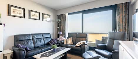 A modern living room with a black leather sofa, a matching armchair, and a small table. There are framed pictures on the wall, a floor lamp, and a large window with curtains.