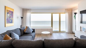 Ett modernt vardagsrum med en grå soffa, ett runt soffbord och ett stort fönster med utsikt över havet.