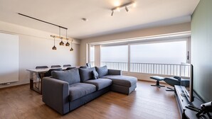 A modern living room with a grey sectional sofa, a dining table with chairs, a TV, and a balcony with a railing. - Apartment in Blankenberge with Sea View (Blankenberge)