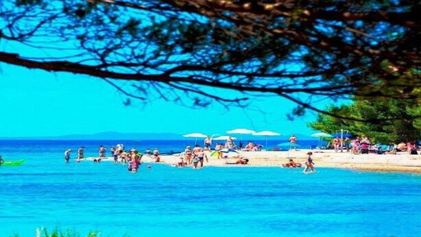 Ein Strand mit badenden und sonnenden Personen, Sonnenschirmen und einem strahlend blauen Himmel.