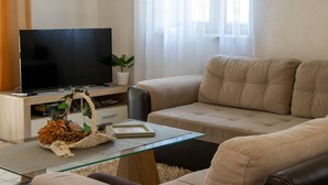 Ein Wohnzimmer mit einem beigefarbenen Sofa, einem Glastisch, einem Fernseher auf einem Holzständer und einer Zimmerpflanze.