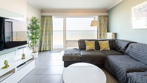 A modern living room with a flat-screen TV, a sofa, a coffee table, and a large window with curtains. - Apartment in Blankenberge with Sea Views (Blankenberge)