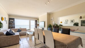 En moderne stue med sofa, spisebord og stole. Et fjernsyn er monteret på væggen, og der er forskellige dekorative elementer.