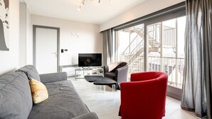 En moderne stue med en grå sofa, en rød lænestol, et fjernsyn og en balkon med udsigt over byen.