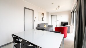 En moderne stue med et hvidt spisebord, sorte stole, en grå sofa og en rød lænestol. Der er et fladskærms-tv og et maleri på væggen.