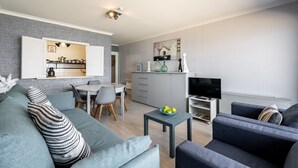 Uma sala de estar moderna com um sofá cinza, uma pequena mesa com frutas e uma TV. Uma cozinha é visível ao fundo, com uma área de refeições.