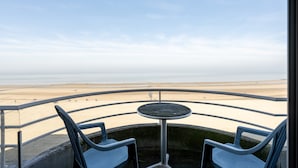 Ein Balkon mit rundem Tisch und zwei Stühlen, der Blick auf einen Strand mit blauem Himmel gewährt.