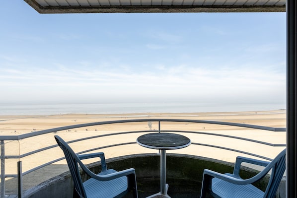 A balcony with a round table and two chairs, overlooking a beach with a clear sky.