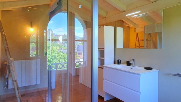 Ein modernes Badezimmer mit einem großen Spiegel, weißen Schränken und Blick durch eine Glastür auf einen Balkon.