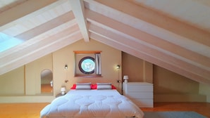 Ein Schlafzimmer mit schrägen Wänden, einem Bett mit weißer Bettwäsche, einem runden Spiegel und einem kleinen Nachttisch mit Lampe.