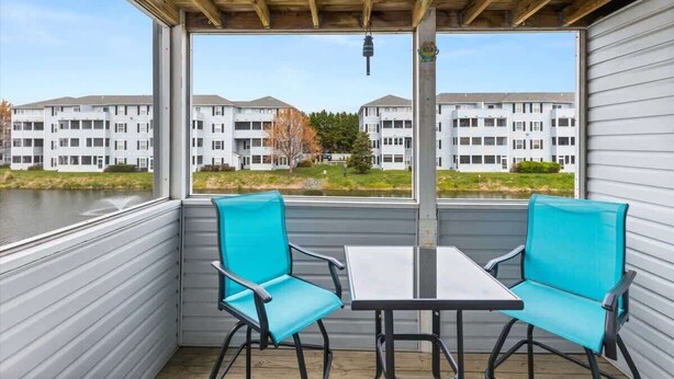 A covered outdoor seating area with two turquoise chairs and a table, overlooking a body of water and a row of white apartment buildings. - Sanibel Summer Hideaway | Waterfront Oasis (Rehoboth Beach)