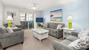 A living room with grey sofas, a white coffee table, and a TV. There is a painting of a beach scene on the wall. - Sanibel Summer Hideaway | Waterfront Oasis (Rehoboth Beach)