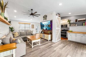 A modern living room with a sofa, coffee table, and TV. There is a kitchen area with white cabinets and a wooden countertop.