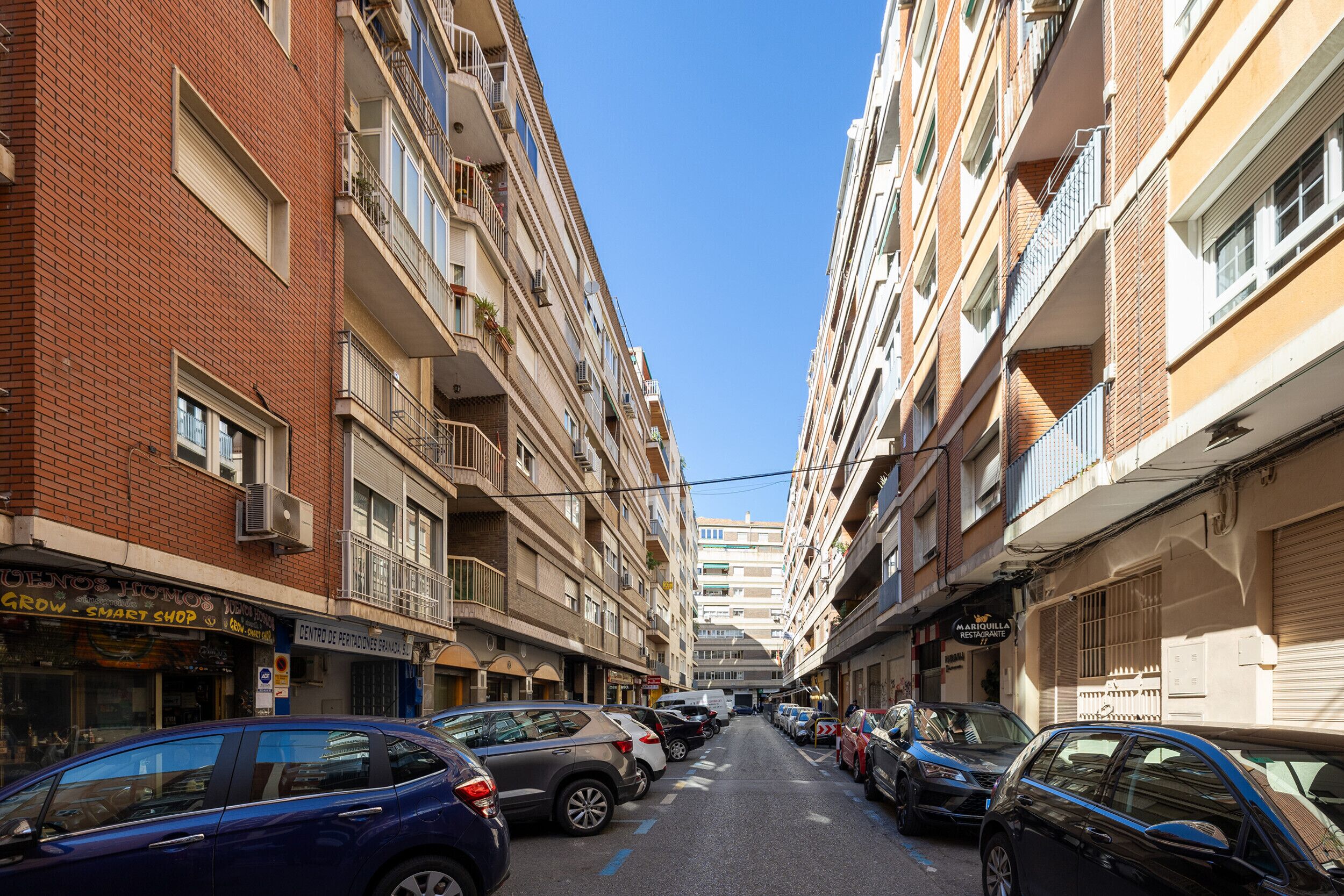 A narrow urban street flanked by multi-story buildings with balconies, parked cars, and various storefronts.