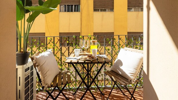 A balcony with a table set for two, a potted plant, and a view of a yellow building. - ChezmoiHomes Gran Vía 47 (Granada)