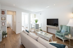 A modern living room with a sofa, coffee table, and a television. There is a dining area with chairs and a table in the background.