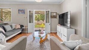 Un salon moderne avec une table basse en verre, des canapés gris, un téléviseur et une vue sur un jardin, visible à travers une porte ouverte.