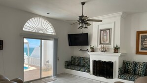 Un soggiorno con ventilatore a soffitto, camino, televisione e vista sull'oceano tramite una porta in vetro.