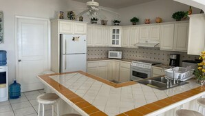 Una cucina con mobili bianchi, un frigorifero, un forno a microonde, un forno, un lavello e una ventola a soffitto. Sono presenti due sgabelli e un dispenser d'acqua.